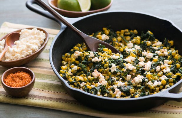 Una sartén con maíz, espinacas y queso Cotija sobre una servilleta junto a tazones de queso y pimienta de cayena.